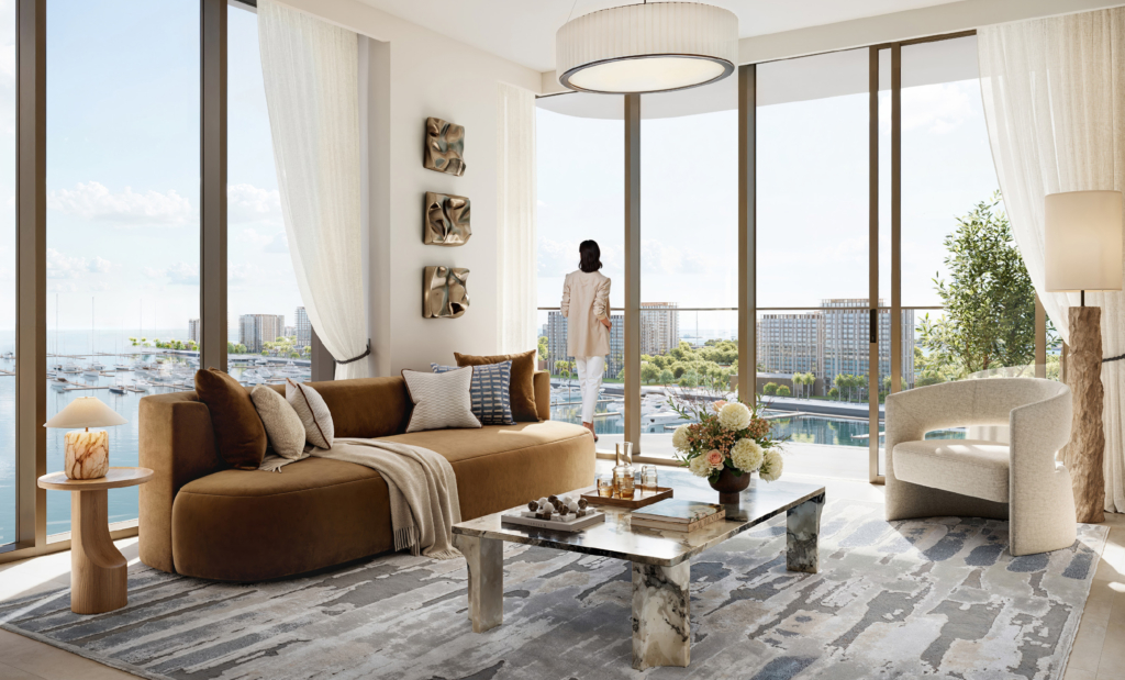 Modernes Wohnzimmer der Baystar by Vida Residenzen in Dubai mit raumhohen Fenstern und Panoramablick auf die Marina. Hochwertige Einrichtung in warmen Naturtönen, Designer-Sofa, Marmor-Couchtisch, stilvolle Dekoration und eine Frau auf dem Balkon schaffen eine luxuriöse Wohnatmosphäre.